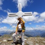Ulrike Wanner machte sich auf ins Neuland: Sie steht vor einem Gipfelkreuz der Kreuzeckgruppe. Blauer Himmel, Quellwolken, Gebirgsketten der Ostalpen im Hintergrund.