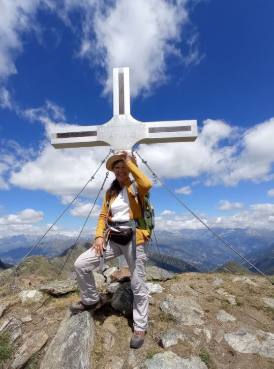 Ulrike Wanner machte sich auf ins Neuland: Sie steht vor einem Gipfelkreuz der Kreuzeckgruppe. Blauer Himmel, Quellwolken, Gebirgsketten der Ostalpen im Hintergrund.