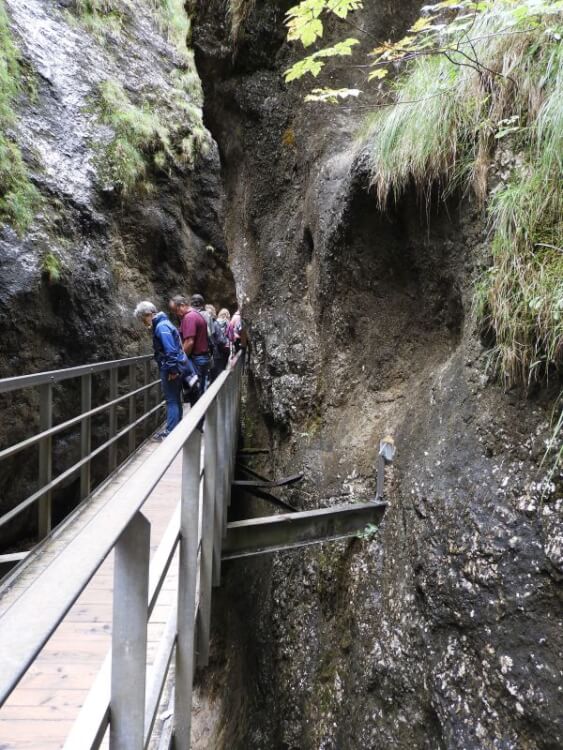Steg mit Metallgeländer an einer steilen Felswand in der Almbachklamm, Wanderer blicken nach unten