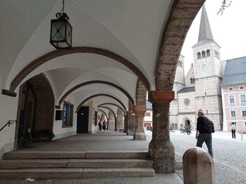 Gewölbter Arkadengang an einem Platz, rechts ein Kirchturm mit Uhr