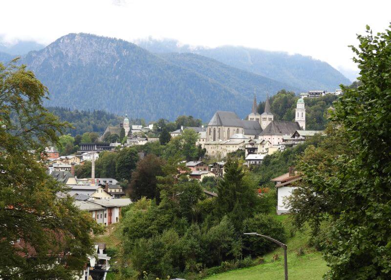 Blick über Berchtesgadens Dächer und Bäume auf eine Kirche mit zwei Türmen vor bewaldeten Bergen