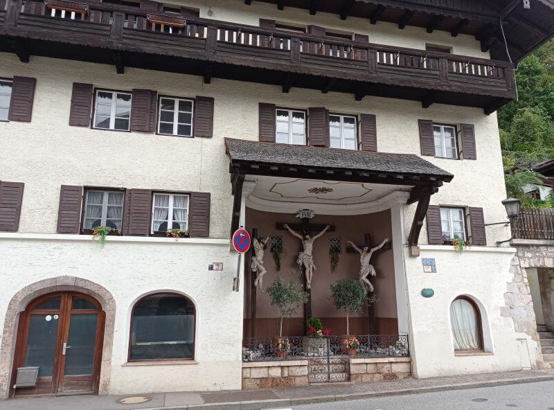 Hausfassade mit Kreuzigungsgruppe unter einem kleinen Vordach, dunkle Fensterläden und Holzbalkon