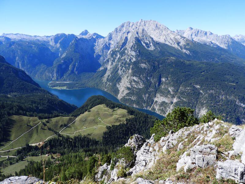 Blick vom Jenner auf den blauen Königssee zwischen steilen Bergwänden, davor eine Almwiese mit Wegen.