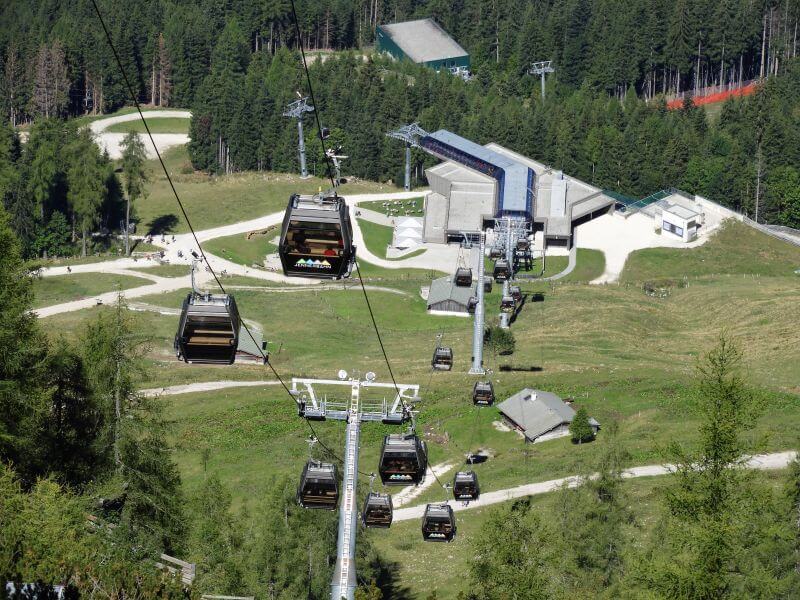 Gondeln der Seilbahn schweben über Wald und Almwiesen zur Mittelstation.