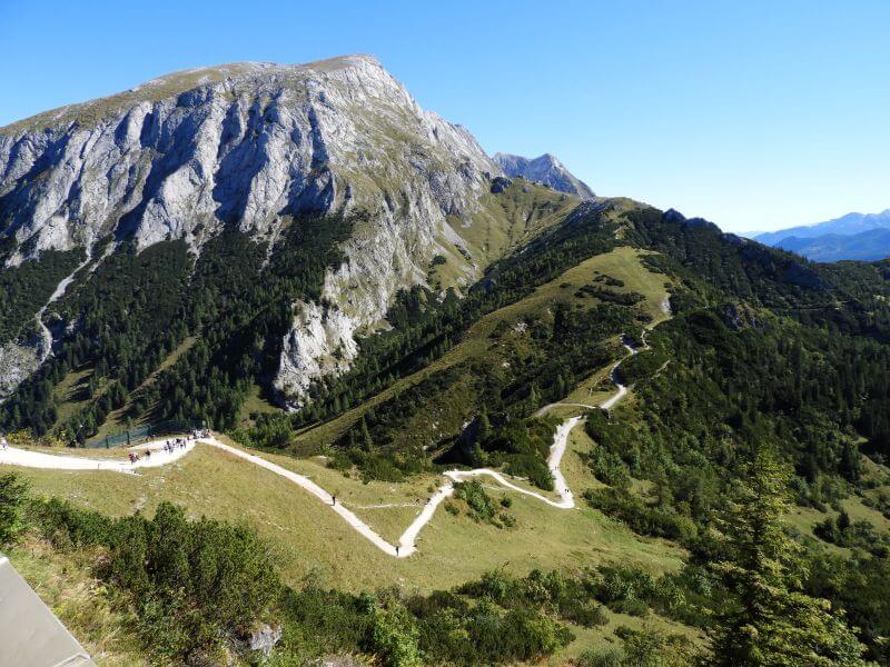 Heller Wanderweg schlängelt sich in Serpentinen über eine Almwiese zu einem felsigen Berg.