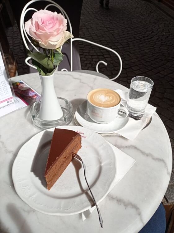 Stück Sachertorte und Cappuccino mit Herz im Milchschaum auf einem Marmortisch, daneben eine rosa Rose