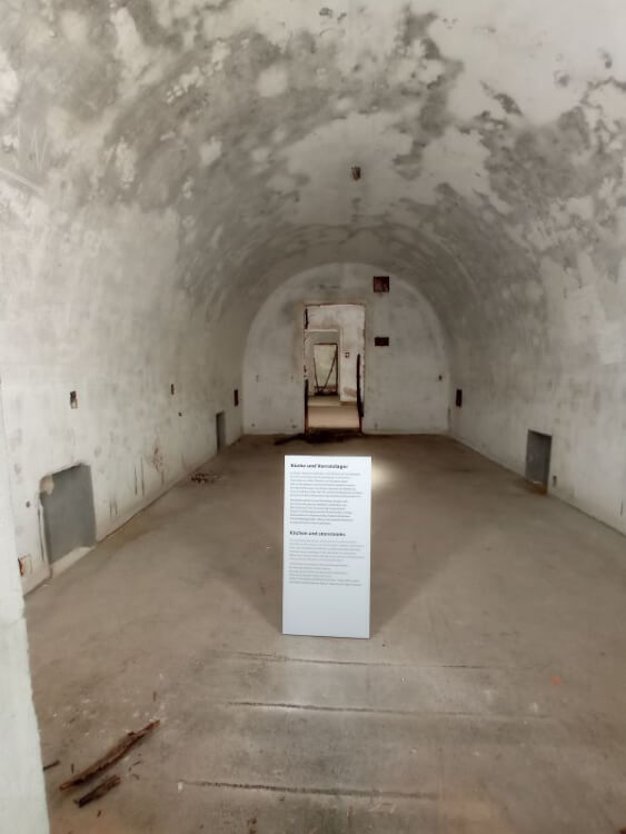 Leerer gewölbter Betonraum im Bunker am Obersalzberg mit Infotafel Küche und Vorratslager, im Hintergrund eine offene Tür