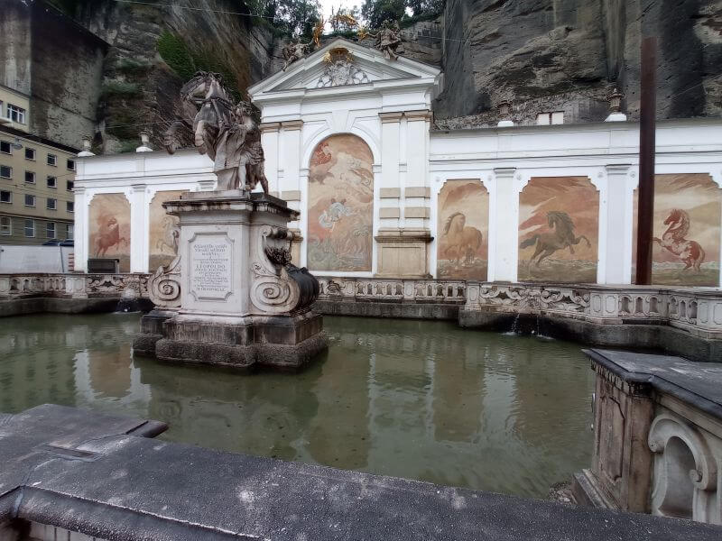 Pferdeschwemme in Salzburg: Wasserbecken mit Pferdestatue vor weißer Wand mit Pferdefresken und Felswand
