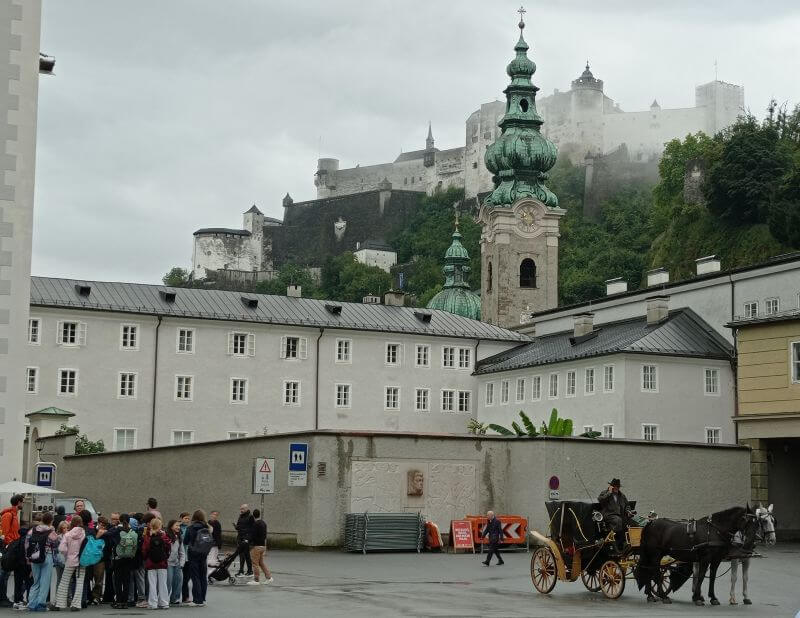 Kirchturm mit grüner Zwiebelhaube, darüber die Festung Hohensalzburg im Dunst, davor eine Pferdekutsche und eine Gruppe Touristen