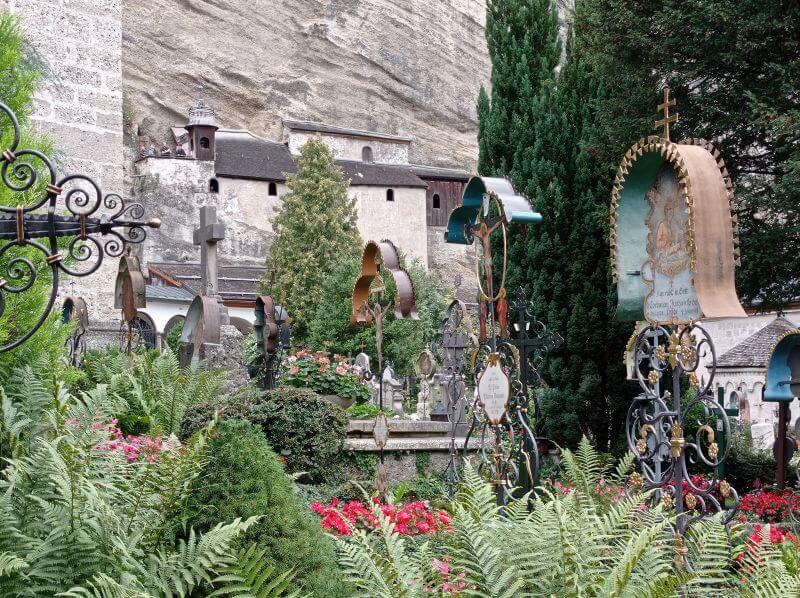 Schmiedeeiserne Grabkreuze und Blumenbeete vor einer Felswand mit Kapellen auf dem Petersfriedhof
