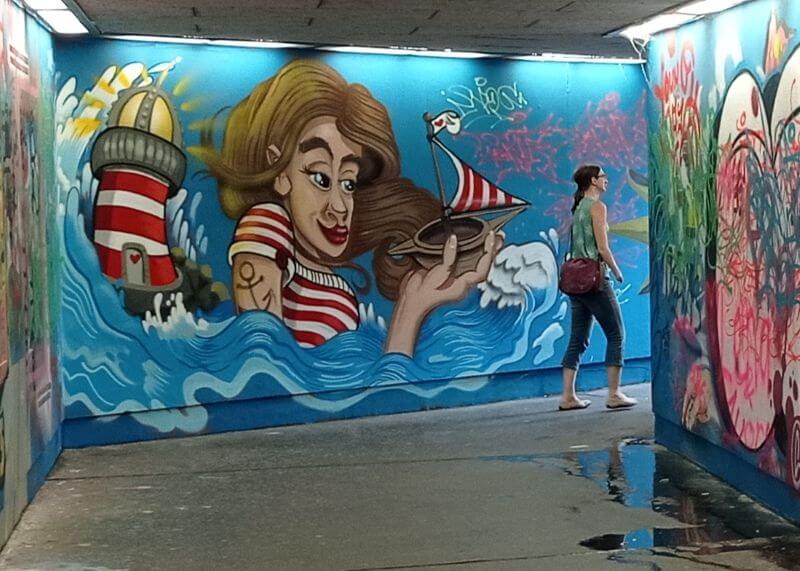 Wandbild in einer Unterführung: Frau im Ringelshirt hält ein Segelboot im Meer, daneben ein Leuchtturm