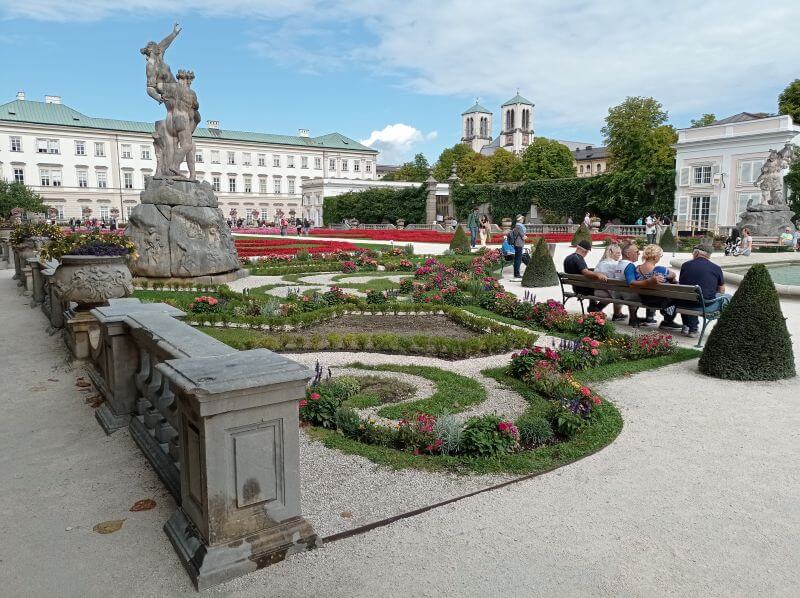 Mirabellgarten mit Blumenbeeten, Statuen und Besuchern auf einer Bank vor dem Schloss