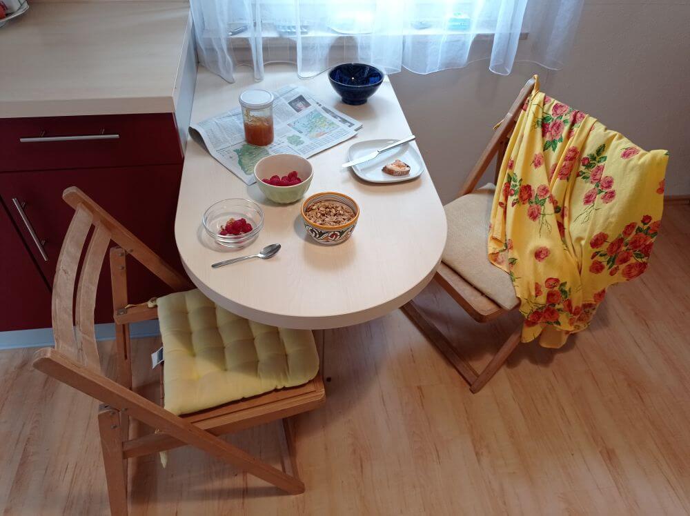 Frühstückstisch mit Zeitung, Himbeeren und Müsli, über der Stuhllehne hängt ein gelbes Sommerkleid mit roten Rosen