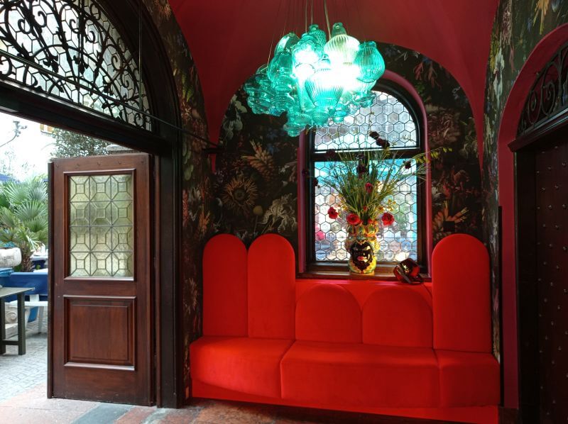 Rote Sitzbank in einer Nische mit gemusterter Wand, grünem Glas-Lüster und Blumenvase vor dem Fenster