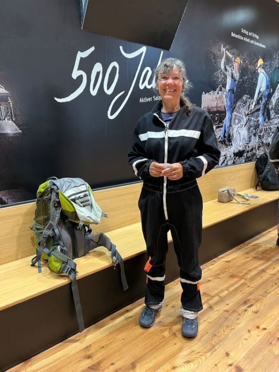 Ulrike Wanner im schwarzen Overall mit Reflexstreifen vor einer Wand mit Bergleuten, daneben ein grüner Rucksack
