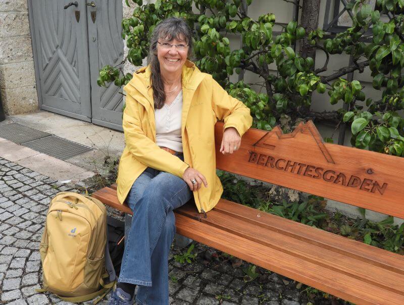 Ulrike Wanner in gelber Regenjacke auf einer Holzbank mit der Aufschrift Berchtesgaden, daneben ein gelber Rucksack